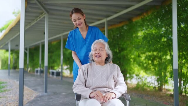 Smiling caregiver pushes elderly woman in wheelchair. Outdoor path with trees and covered walkway. Caregiver's gentle smile shows kindness