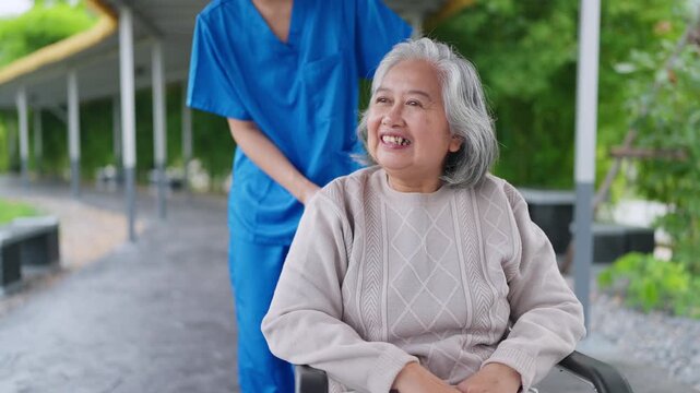 Smiling senior in wheelchair, outdoors. Caregiver in blue uniform beside her. Woman gestures to something in distance. Senior smiles warmly