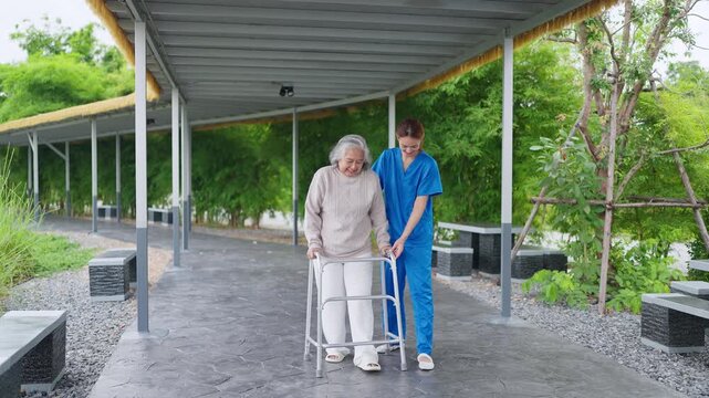 Senior woman uses walker with care. Caring nurse walks beside her. They smile under sheltered path. Green trees frame the scene