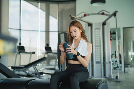 Young woman athlete sitting on exercise equipment holding water bottle taking break in modern gym with natural light