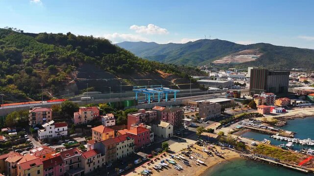 Aerial view of Vado Ligure, Italy, showing a beachside town, marina, industrial port, highway and rail infrastructure between turquoise water and green Ligurian hills.