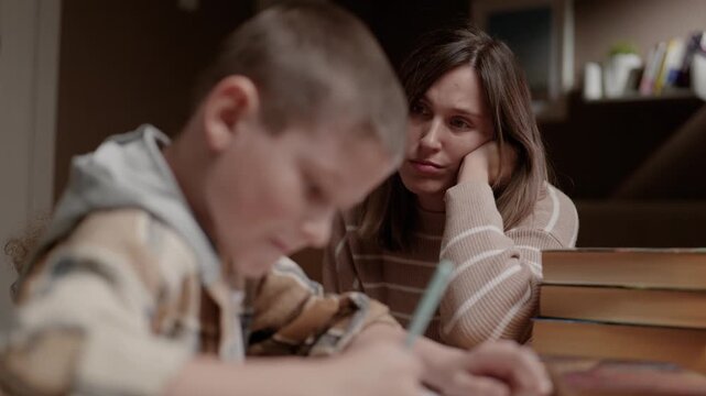 Schoolboy doing homework while mother sits beside him and observes. Parenting education supervision family concept
