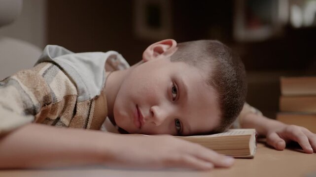 Tired exhausted schoolboy lying face down on book and looking sadly at camera. Fatigue homework learning stress childhood concept