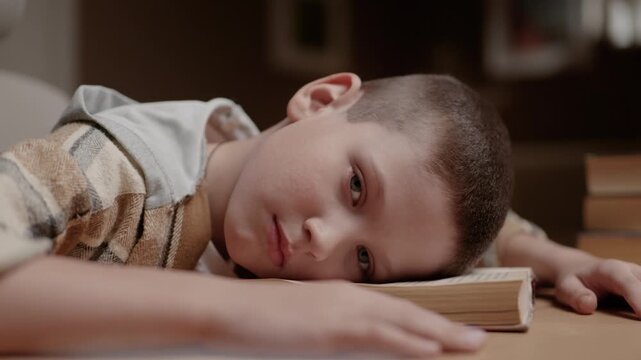 Exhausted schoolboy lying face down on book and sadly looking into camera as it slowly zooms in. Fatigue homework learning stress childhood concept