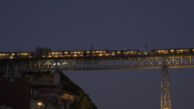 Illuminated metro trains pass across the upper deck of Lu&iacute;s I Bridge in Porto, above hillside buildings in the evening.