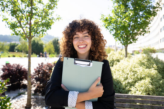 Smiling white woman holding folder sitting on bench in outdoor park