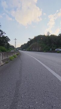 Vertical footage of a winding asphalt road beside a forested hill, with concrete roadside barriers, streetlights and soft golden clouds in the evening sky.