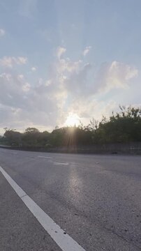 Vertical footage of golden evening sunlight shining through roadside trees above an asphalt road, with a concrete barrier, forested greenery and soft clouds in the sky.