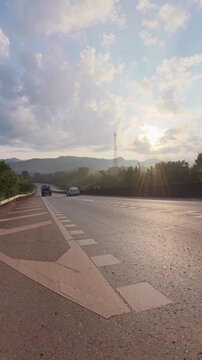 Vertical footage of a mountain road descending toward a distant valley, with passing vehicles, layered forested hills and dramatic clouds illuminated by warm evening sunlight.