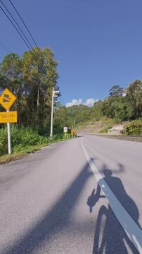 Vertical downhill cycling POV on a sunny road curving through green forested hills, with the cyclist and bicycle casting a long shadow across the pavement beneath a clear blue sky.