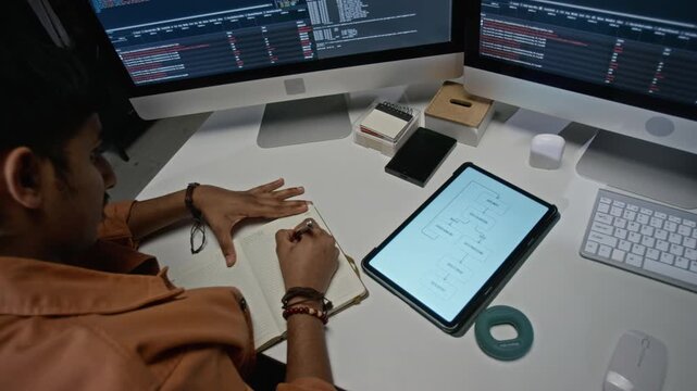 Young adult male developer taking notes in notebook near tablet displaying flowchart while sitting before multiple computer monitors. Ideal for software engineering corporate campaigns