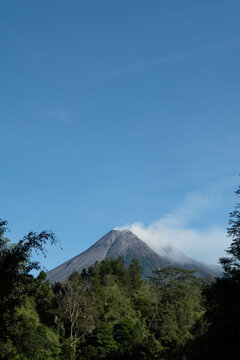 Mount Merapi stands tall against the sky with a small white smoke plume, an eroded volcanic crater and lush green vegetation surrounding the mountain