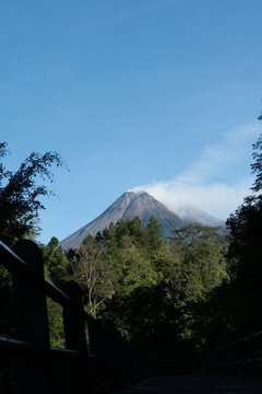 Vertical landscape of Mount Merapi's rugged volcanic peak with faint white smoke drifting upward, surrounded by tropical greenery and terrain scarred by past eruptions
