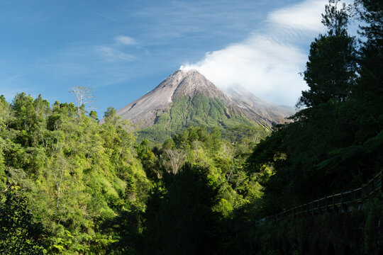 Mount Merapi active volcano in Yogyakarta, Indonesia, with a thin plume of white smoke rising above its rugged crater and lush green vegetation covering the surrounding slopes