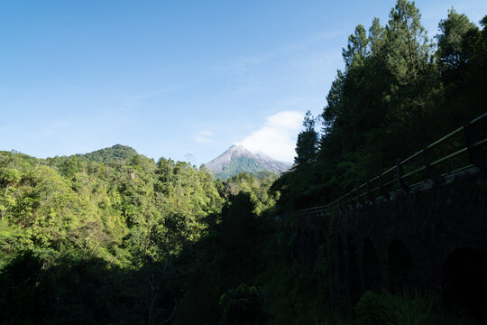 Scenic view of Mount Merapi with white volcanic smoke above its weathered summit, surrounded by green hills and rugged terrain shaped by past eruptions