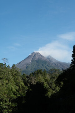 Mount Merapi emerging through morning mist, with its dark volcanic silhouette rising above a sea of clouds and faint white smoke drifting from the summit