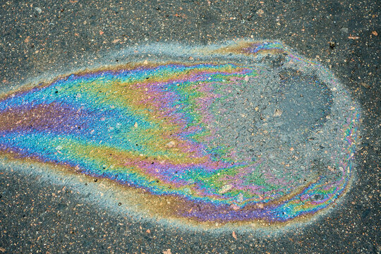 Colorful oil slick rests on rain darkened asphalt, with rainbow film outlining shallow water and scattered grit. Vivid iridescent pattern offers abstract view of urban road surface pollution.