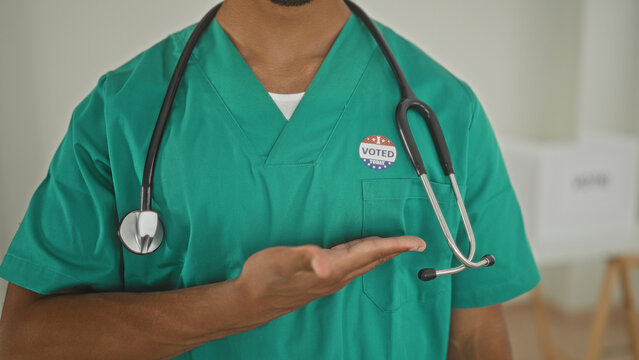 Man doctor in green scrubs points finger to voted sticker while wearing stethoscope inside building; pride civic duty.