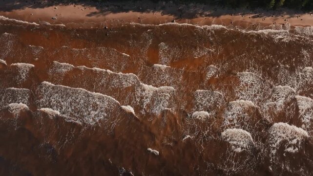 Rows of brown foaming waves breaking near beach from above. Drone shot of rough muddy surf with white crests and beach edge lit by low autumn sun.