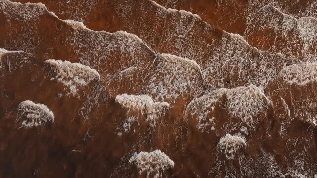 Brown sea surf and white foam texture from directly above. Close drone shot of rough muddy water, breaking wave crests and swirling white foam patterns.