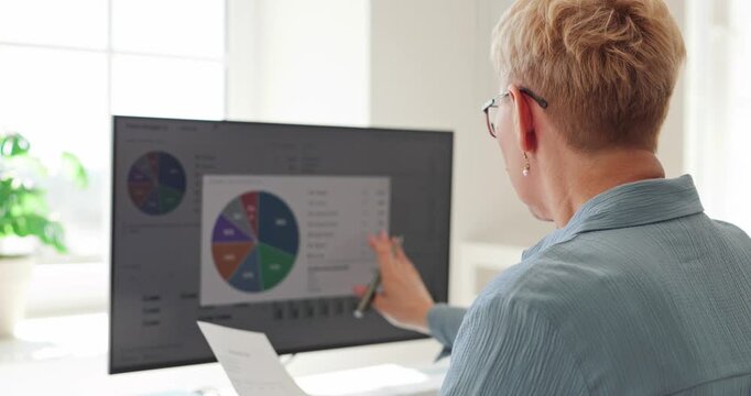 Mature woman analyzes budget charts and bills. She uses a computer dashboard to review expenses and finance data for monthly household planning. Everyday digital money management concept.