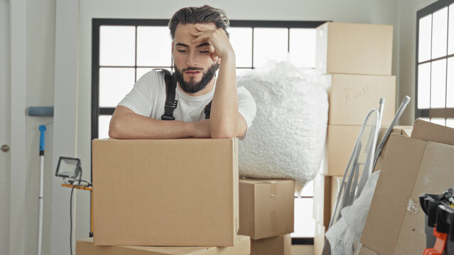 Man mover wearing apron leans on stacked cardboard boxes amid bubble wrap and packing materials, hand on forehead in building; weary patience.