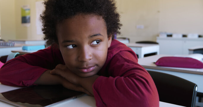 Bored african american schoolgirl leaning on desk looking to window in elementary school classroom