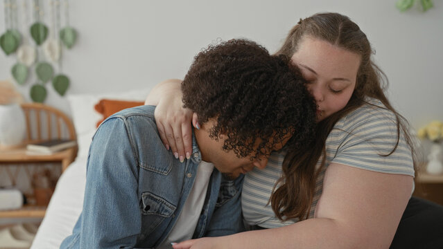 Plus size woman embraces man, hand on his shoulder and arm around his back as he leans in with head bowed on her shoulder on bed inside a building; comfort support.