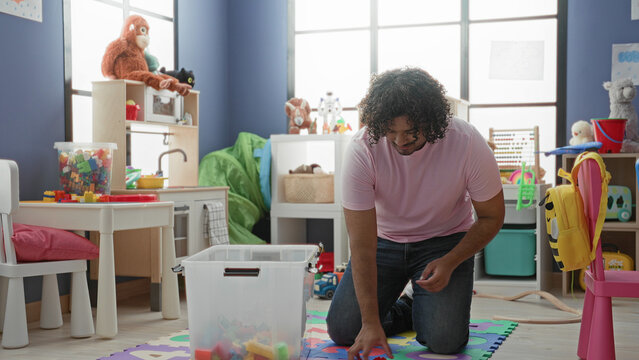 Man kneels and picks up toy blocks from a clear plastic bin in a kindergarten playroom building, smiling while arranging pieces; nurturing.