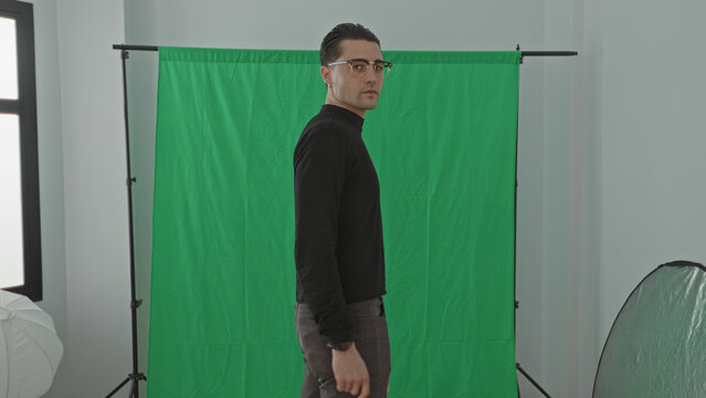 Man in black sweater with hand at waist standing before a green screen backdrop in a studio turning his torso and looking over his shoulder while taking a step; calm.