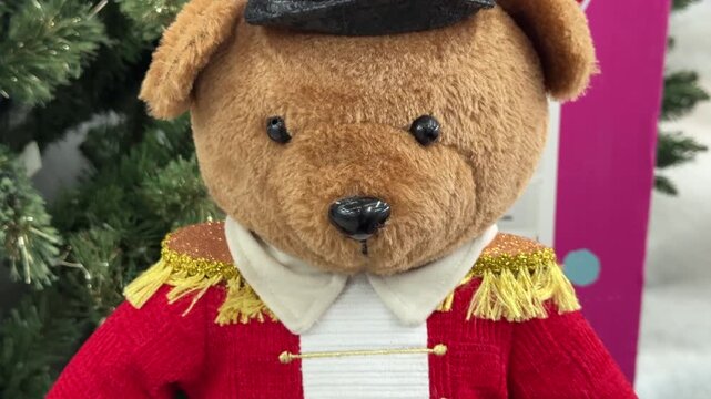A brown teddy bear wears a red toy soldier uniform with gold fringed epaulettes and a tall decorative hat beside an artificial Christmas tree.