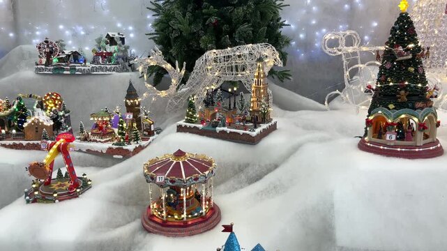 A miniature carousel turns among illuminated Christmas village models, decorated trees and wire reindeer arranged on artificial snow.
