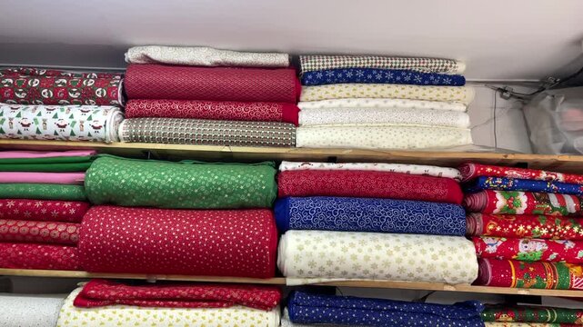 Folded and rolled fabrics fill wooden shelves, displaying Christmas characters, snowflakes, stars and scrolling patterns in contrasting colors.