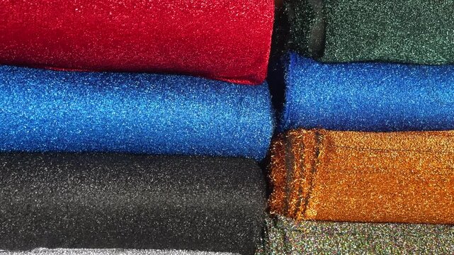 Folded glitter fabrics sit in two adjacent stacks, with blue, gold, black and silver textiles and a multicolored iridescent layer visible in close-up.