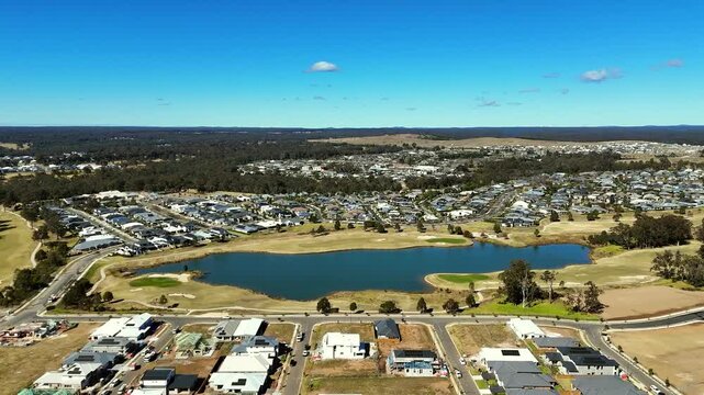 Wilton houses and vacant plots surround a lake bordered by golf course greens and trees, aerial establishing