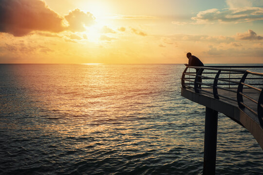 Peaceful sunset by sea with solitary man on pier enjoying golden hour, coastal travel and quiet contemplation, serene ocean horizon and warm evening sunlight