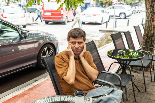 Noise pollution and urban stress affecting man covering ears at street cafe, traffic noise causing discomfort and sensory overload in busy city environment