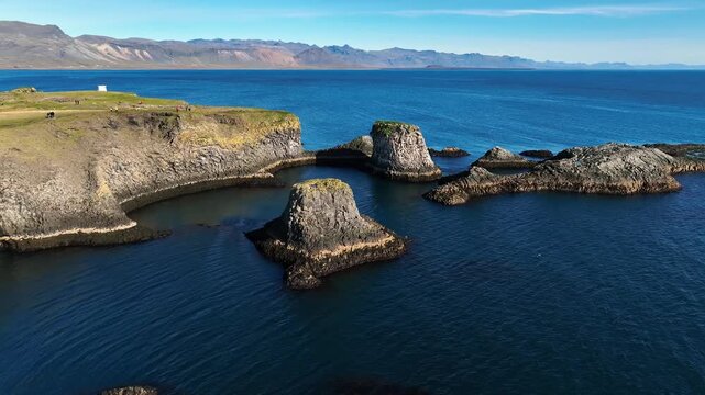 Drone view of volcanic sea stacks off Arnarstapi, Iceland, surrounded by deep blue Atlantic water and rugged basalt cliffs beneath distant mountains.