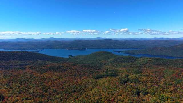 Sweeping panorama from Sleeping Beauty Mountain overlooks Lake George, fiery autumn canopy spreading toward Adirondack ridgelines beneath blue sky with scattered clouds. Aerial drone wide shot