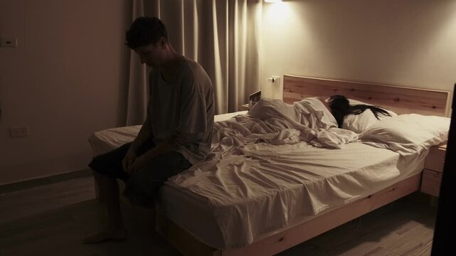 Millennial couple experiencing emotional distance in an everyday bedroom scene, woman focused on her laptop while a sad man sits on the bed looking down, relationship problems and emotional isolation.