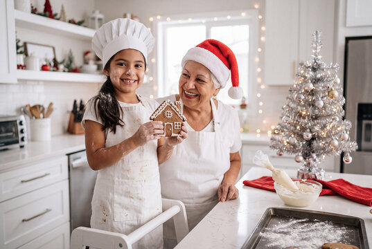 Abuela con gorro de papa noel y nieta con gorro y mandil de chef manchada de harina, ense&ntilde;ando galletas de jengibre en forma de casita que acaban de hacer juntas, sobre fondo de coocina blanca