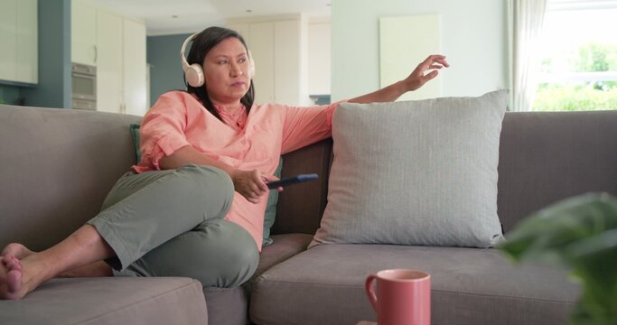 Mature adult woman sitting on gray sofa in living room, wearing headphones, holding TV remote