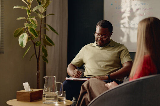 African American male psychologist taking notes during therapy session with teenage patient. Useful for mental health counseling services, psychology clinic advertising and youth support campaigns