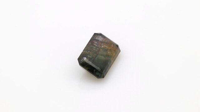 Polished emerald cut polychrome tourmaline gemstone features multiple brilliant geometric facets reflecting bright illumination across a clean neutral surface during smooth rotation.
