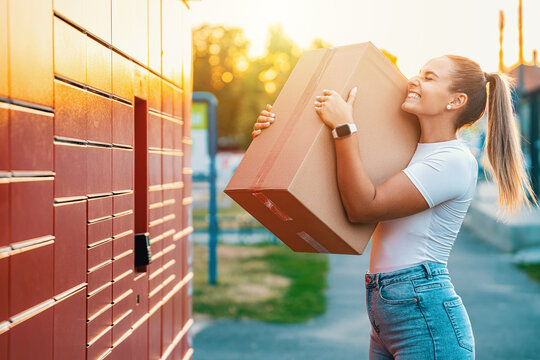Woman carrying a large cardboard parcel beside outdoor delivery lockers in evening light