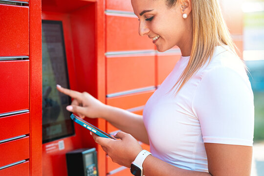 Smiling woman using a parcel locker touchscreen while checking delivery on her smartphone