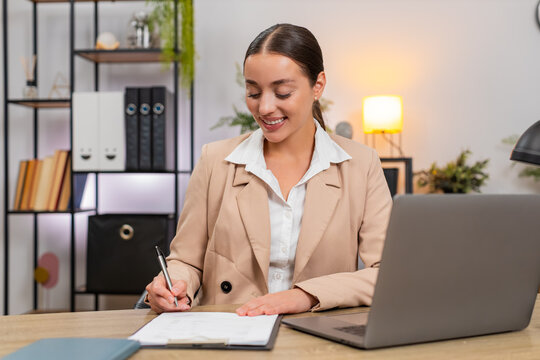 Young businesswoman at home office reads contract on laptop checks clauses and prepares pen to sign. Freelancer girl at table signs at end of page writes name adds date saves copy and stores file