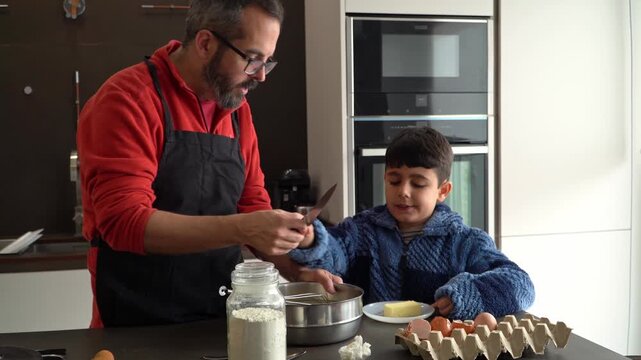Child learning to use a kitchen knife on butter, under the careful supervision of his father.