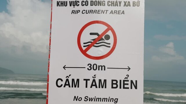 Beach warning sign prohibits swimming within 30 meters of rip current area. Bold bilingual lettering and crossed swimmer symbol stand beside rough surf under cloudy coastal sky, warning swimmers away.