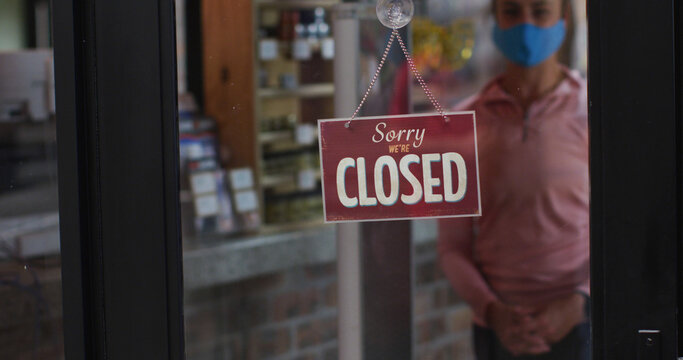 Caucasian female shopkeeper wearing face mask changing closed to open sign in window of sports shop